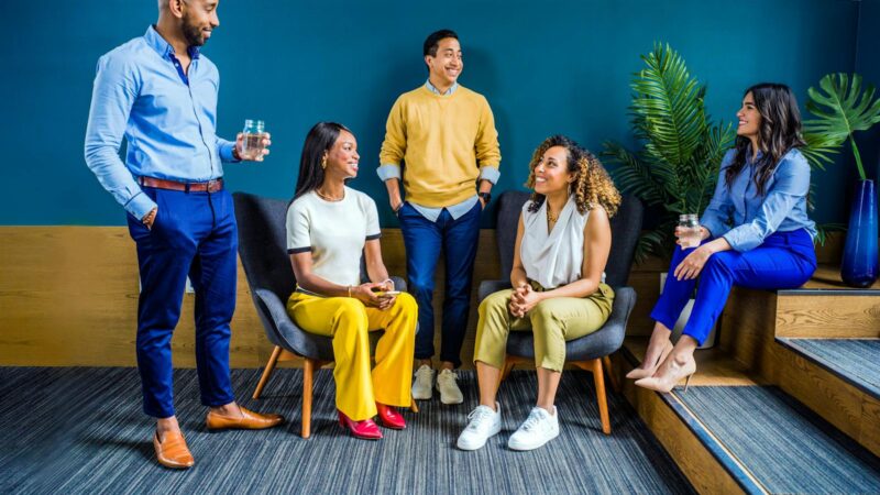 A diverse group of five professionals engaging in a casual indoor meeting, smiling and talking.