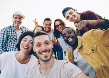 A diverse group of friends smiles while taking a selfie outdoors during the day.